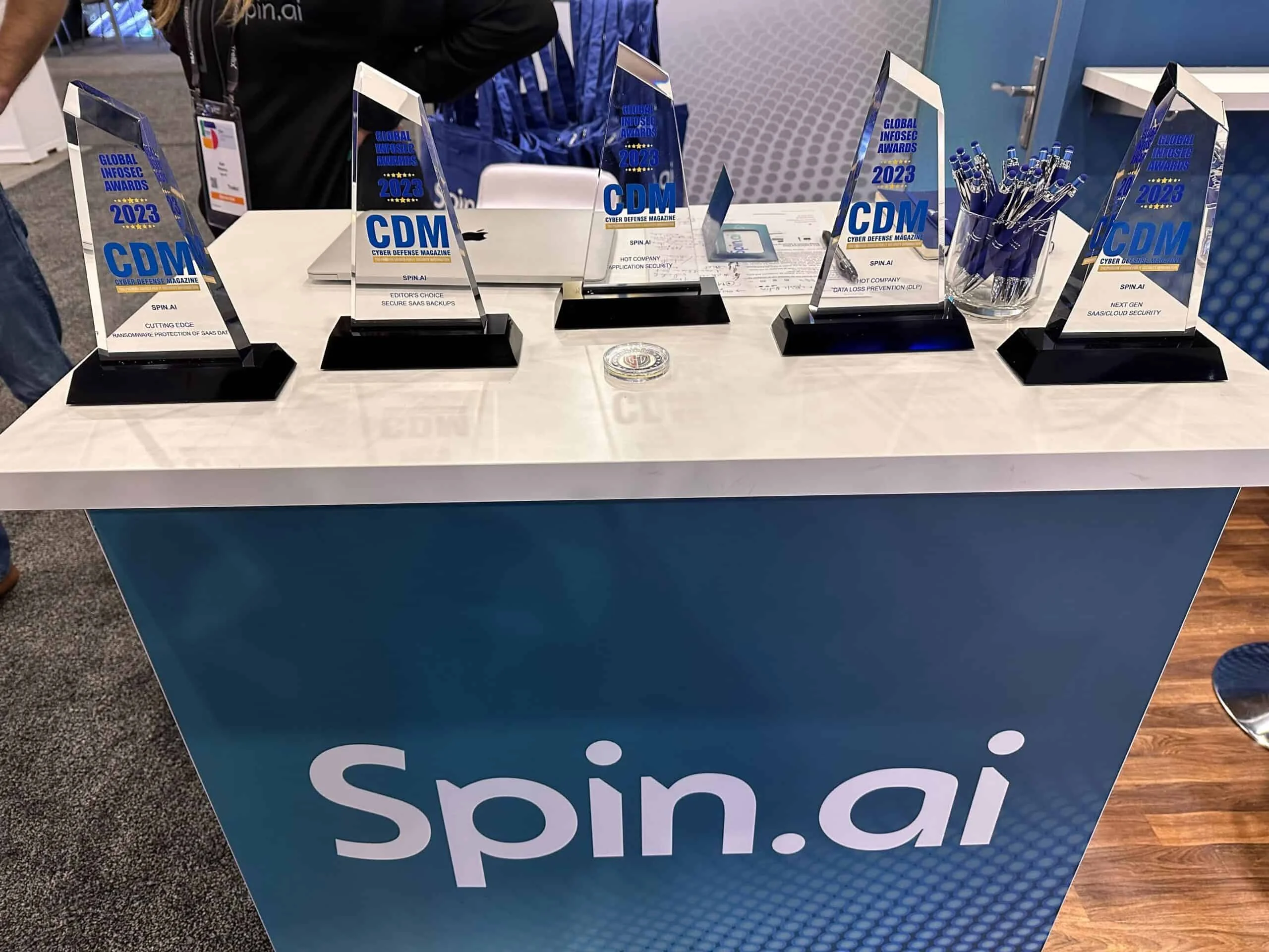 SpinAI at RSA 2023 | Spin.AI Trophy display on a booth table for Spin.ai with clear glass awards reading Global Infosec Awards 2023/2024 and a pen cup nearby.