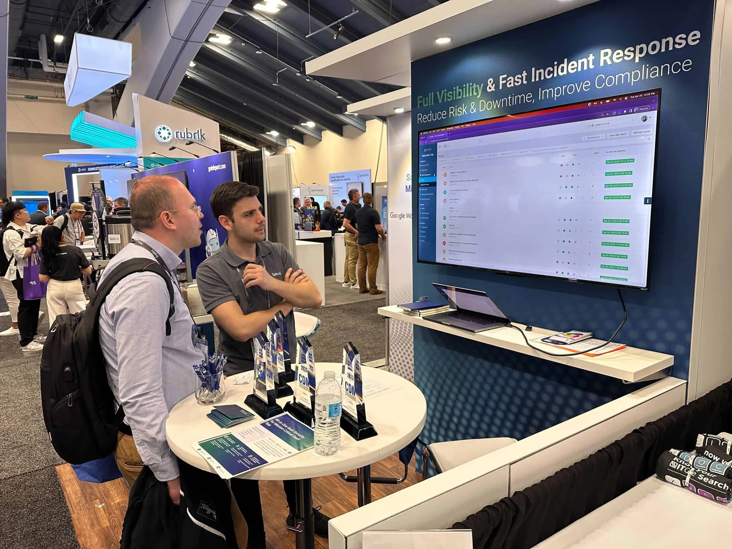 SpinAI RSA 2023 Participation | Spin.AI Two men stand at a booth table with brochures and a laptop, near a large monitor displaying a software dashboard at a trade show.