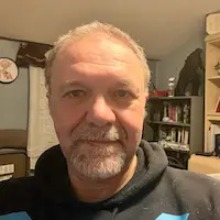 Close-up portrait of a middle-aged man with gray hair and beard wearing a dark hoodie, indoors in a home setting.