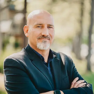 Professional headshot of a bald man with a gray beard wearing a dark blazer, standing outdoors with arms crossed.